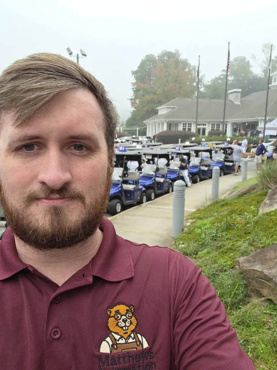 Mike Matthews in a maroon Matthews Automation polo, standing beside the cart line outside the clubhouse.