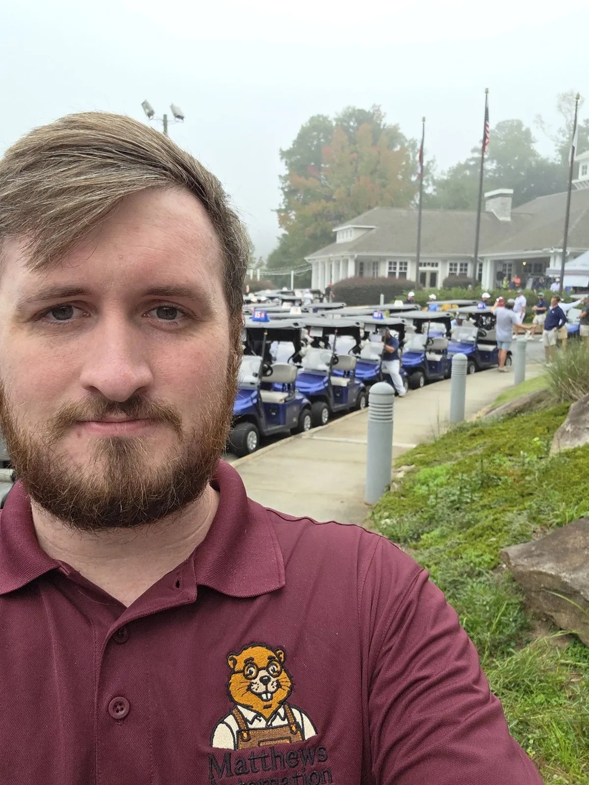 Mike Matthews in a maroon Matthews Automation polo, standing beside the cart line outside the clubhouse.